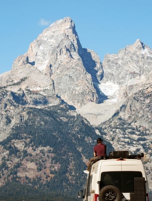 A man sits on the roof of a panel van looking toward a mountain