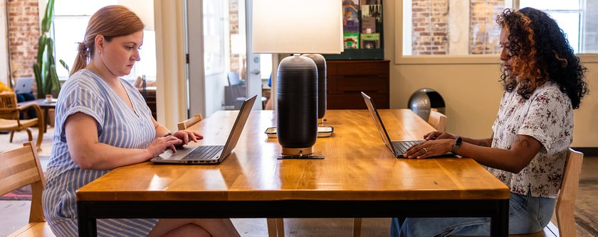 Two women sit at a table facing each other working on their laptops