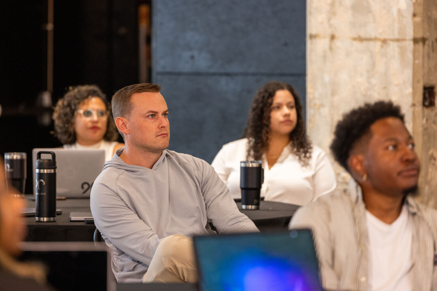 People attentively listening during a meeting, seated at tables with laptops and drinks, against a dark and light background.