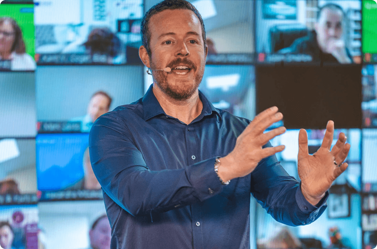 An individual speaking on a stage. Behind him is a group of people in a Zoom meeting watching on.