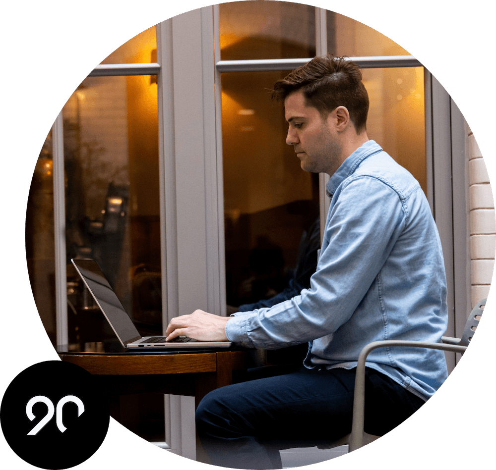 A man seated outside working on his laptop. The Ninety logo is in the corner.